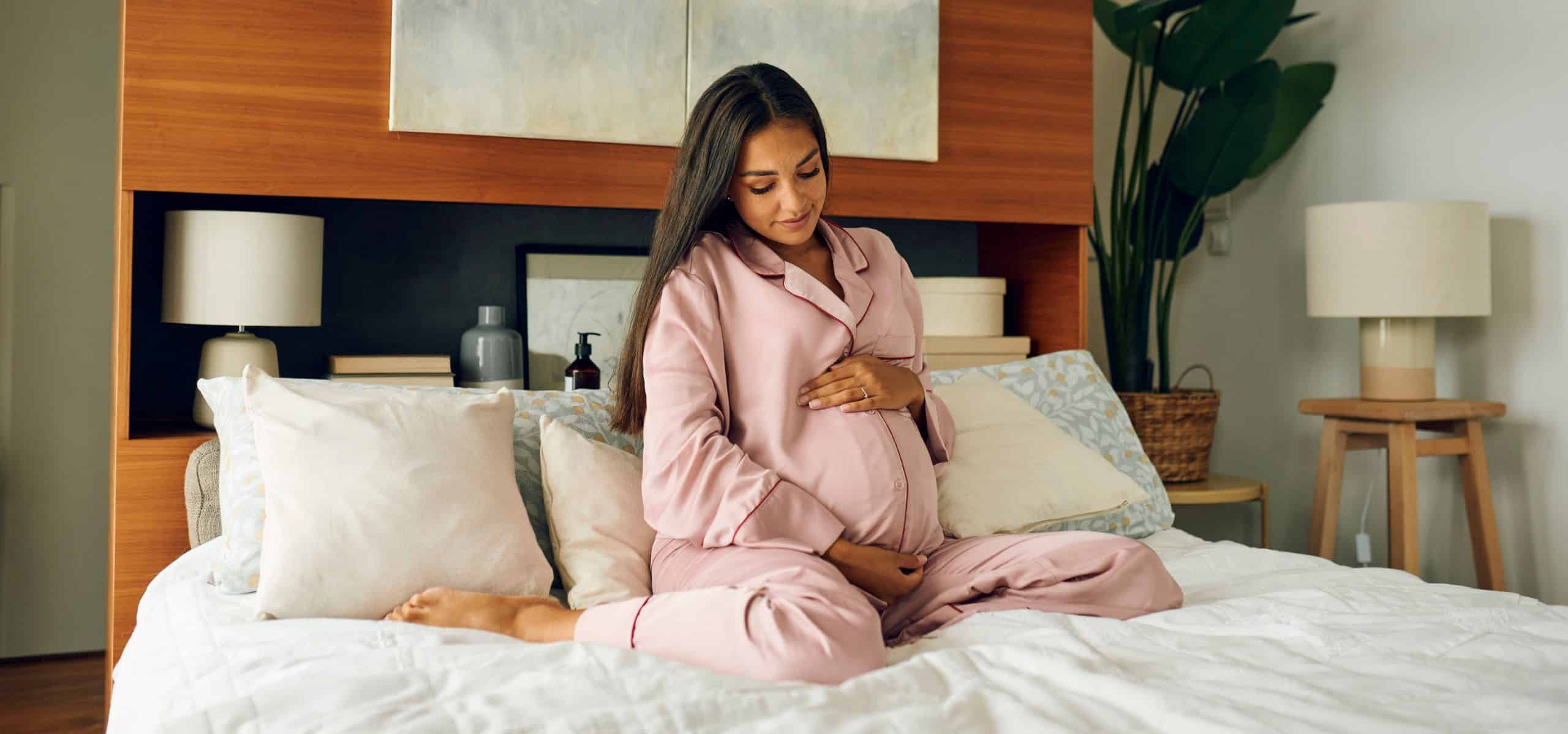 Trudnica u ružičastom ogrtaču sjedi na krevetu i drži se za trbuh.